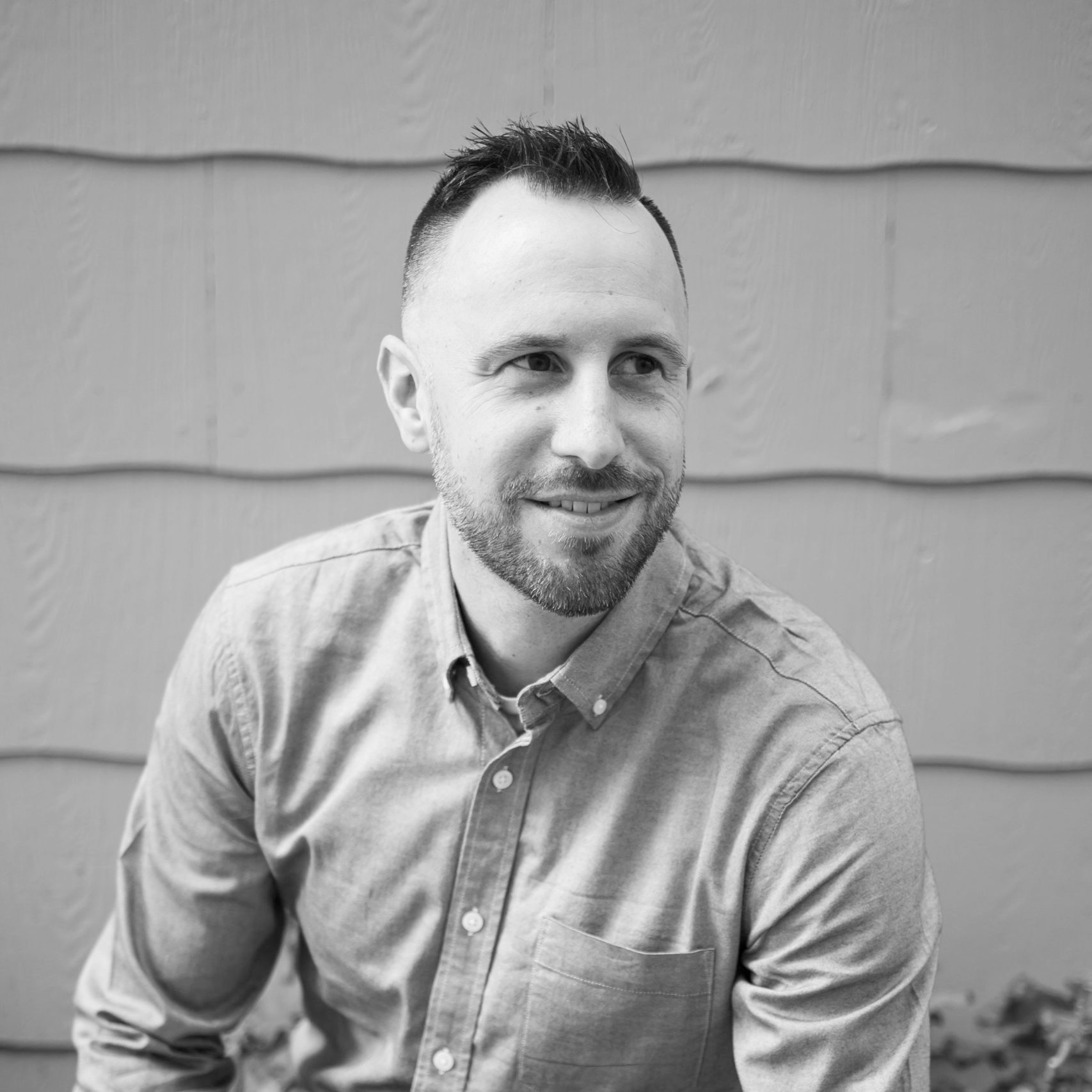 Black and white photo of a smiling man in a button-up shirt, standing in front of a textured wall with horizontal panels.